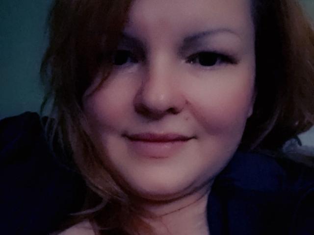 AimeeRocks Gesicht, mit einem leichten Lächeln, heller Haut, braunen, welligen Haaren und dezentem Make-up, trägt ein dunkelblaues Hemd, auf einem unscharfen Foto bei schwachem Licht..