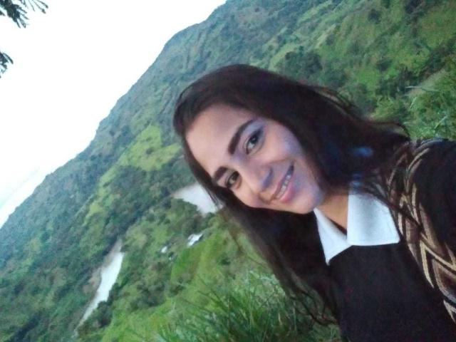 AfroditaHan mit langem, dunklem Haar, braunen Augen, weißer Hemdjacke, schwarzem Pullover, lächelnd, stehend in einer grünen, grasbewachsenen Landschaft, Berge im Hintergrund, süßes Gesicht, kleine Nase, leichtes Lächeln..