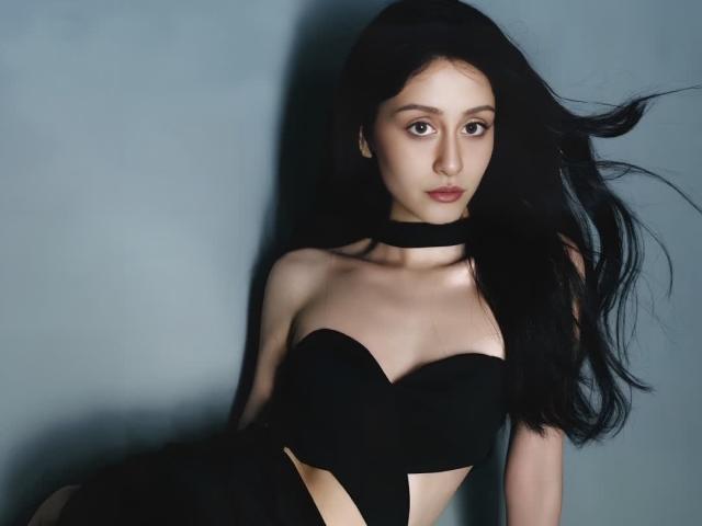 Alesya, com longos cabelos pretos, usando um colar preto e um top preto sem alças, mostrando seus seios e um abdômen plano, fino e pálido, contra um fundo cinza..
