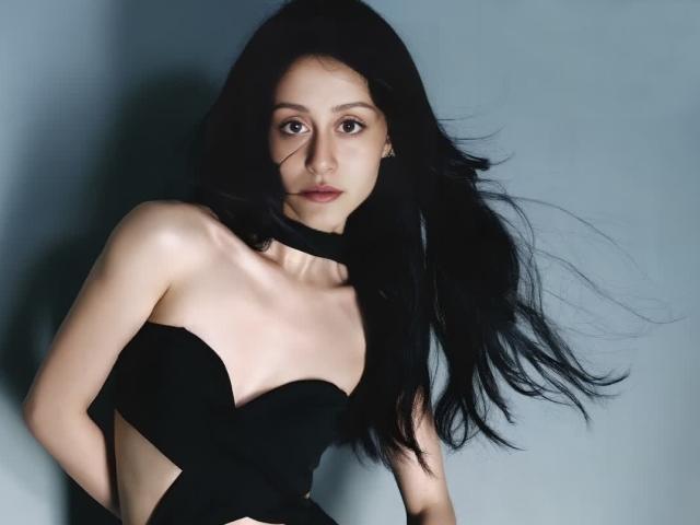AlesyaDrons a les cheveux noirs, porte une robe noire sans bretelles et a une expression sérieuse. On peut voir ses seins et une partie de sa poitrine, sur un fond gris..