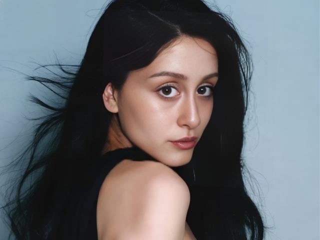Alesya Drons hat langes, schwarzes Haar, dunkle Augen und ein leichtes Lächeln. Sie schaut über ihre Schulter, wobei ihre nackte Schulter sichtbar ist. Sie trägt ein schwarzes Oberteil..