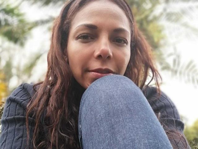 VenusChaud with wavy brown hair, wearing a dark rib-knit sweater, has her knee up, showing blue jeans, smiling slightly, looking down, with a relaxed, natural expression.