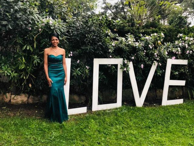 VenusChaud in a green, strapless, satin dress standing on grass, wearing a necklace, next to large white letters spelling "LOVE". She has a cute, slender ass.