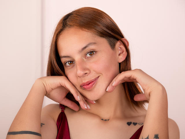 VictoriaOrtega with light brown hair, brown eyes, and a cute smile, resting her chin on her hands, wearing a red strap dress, showing tattoos on her arms and chest.