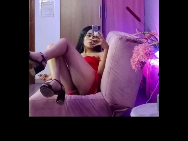SalomeMille with black hair, red dress, and black heels, legs crossed, holding phone, taking a mirror selfie, pink flowers, purple light, wooden door in background.