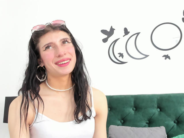 EvelinGonzales heeft lang zwart haar, roze lippenstift, een wit tanktopje, een parelketting, grote oorringen en een zonnebril op haar hoofd, en ze glimlacht met een groen fluwelen bed achter haar..