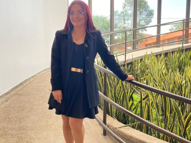EimmyHoskX with red hair, glasses, black dress, black coat, and gold belt, standing by a metal railing, smiling, holding a phone in a sunlit, green, plant-filled area.