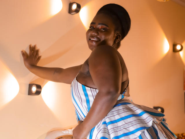 Dilay, dark-skinned with a big smile, wearing a blue and white striped dress, has her back to the camera, showing her ass, with one hand on the wall.