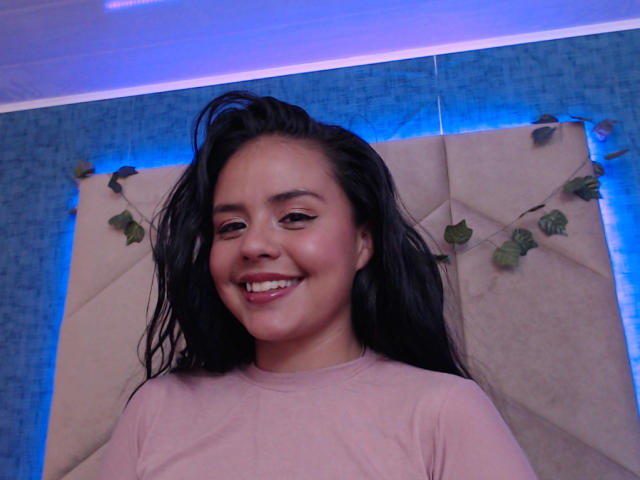 ArielleWilde with wet black hair, smiling with pink lipstick, wearing a pink shirt, blue-lit background, vine decorations, and a beige tufted headboard.