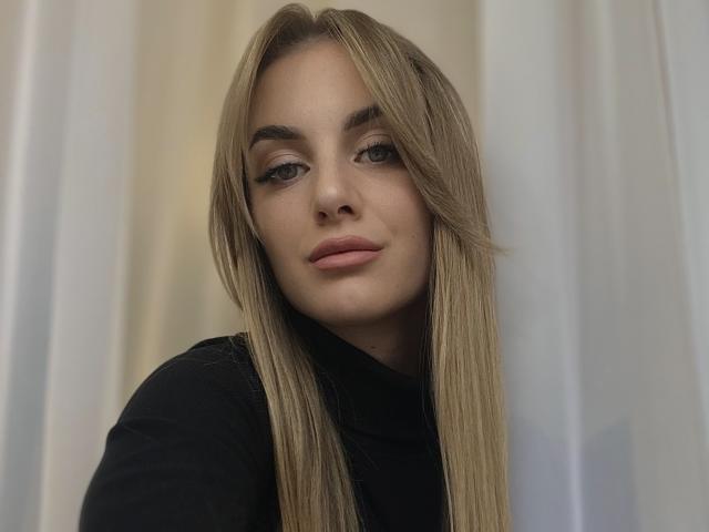 BlondyOneSex ha lunghi capelli biondi lisci, occhi verde chiaro e labbra rosa, indossa una maglietta nera, guarda leggermente di lato con un sorriso sottile e sicuro..