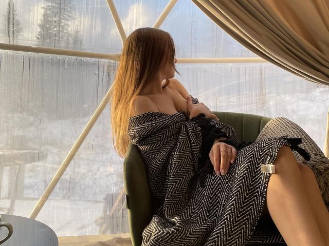 Barbara, con cabello largo y castaño, vestida con un vestido blanco y negro con estampado de zigzag que deja ver sus senos, sentada en una silla verde, con luz solar entrando por la ventana, cortinas beige, nieve afuera, expresión tranquila..