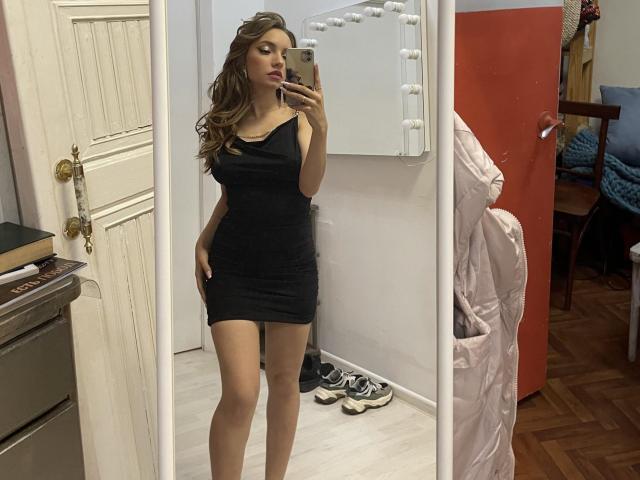 Barbara, avec de longs cheveux bruns, portant une robe noire moulante, montrant ses seins et ses fesses, prend un selfie devant un miroir avec son téléphone, dans une pièce avec des planchers en bois..
