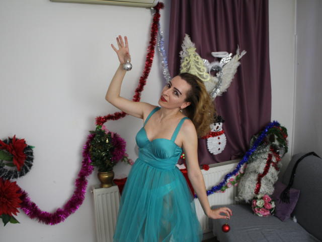 EvelynAngel, con cabello castaño rizado, vestida con un vestido transparente color turquesa, levanta su mano con un anillo de plata, mostrando sus senos, y está de pie en una habitación con decoraciones navideñas..