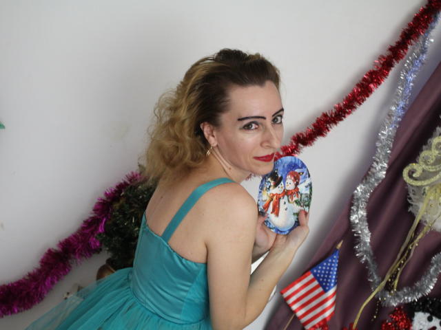 EvelynAngel with curly blonde hair, green dress, holding blue porcelain figurine, looking back, red lipstick, Christmas tinsel, American flag, smiling, medium tits, fair skin, white wall.