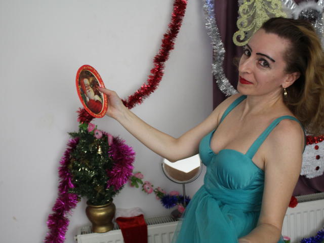 EvelynAngel, avec ses cheveux blonds, son rouge à lèvres rouge, sa robe turquoise, tenant un miroir ovale dans lequel on peut voir ses seins, décorations de Noël, guirlande scintillante, plante, bougie rouge, mur blanc, souriante..