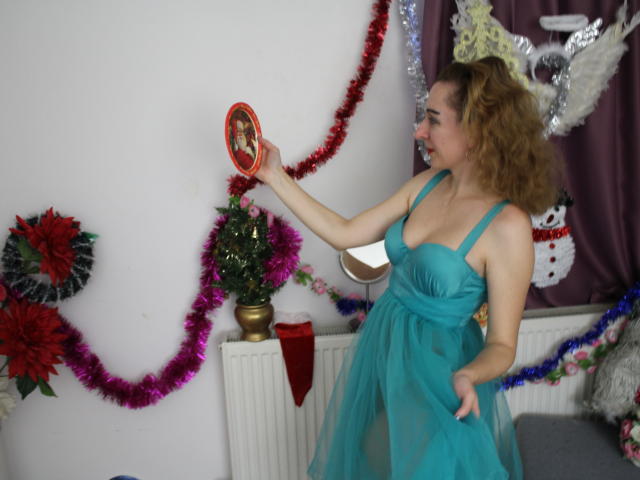 EvelynAngel, vêtue d'une robe bleue, avec des cheveux bruns bouclés, tenant un ornement rouge près d'une décoration de Noël, avec des guirlandes roses et rouges, un petit sapin sur un radiateur, souriante..