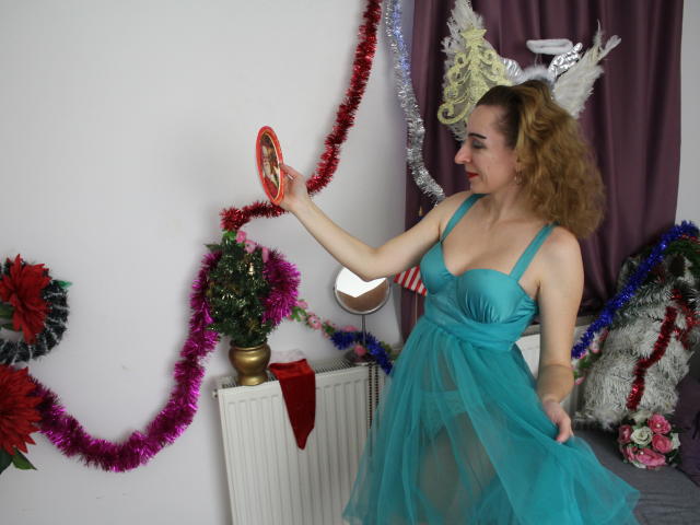 EvelynAngel, met krullend bruin haar, draagt een turquoise, doorschijnende jurk en raakt een ronde spiegel aan. Haar middelgrote borsten zijn zichtbaar. Ze draagt witte hertenhoorns en is omringd door kleurrijke kerstdecoraties..