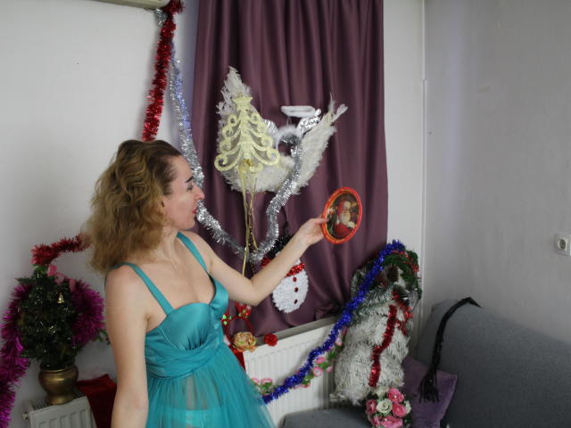 EvelynAngel with curly blonde hair, blue-green dress, holding a white angel ornament, silver tinsel, and red garland, standing in a room with Christmas decorations.