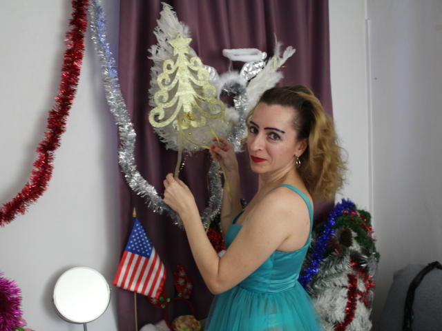 EvelynAngel, avec ses cheveux blonds bouclés, son rouge à lèvres rouge et sa robe bleue, tenant des décorations de Noël argentées et blanches, regardant la caméra, debout devant un rideau violet..