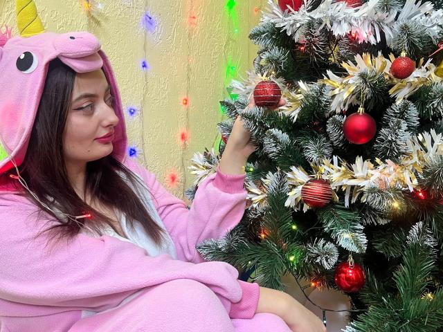 MilanaPrice in pink unicorn onesie with booby-like hood, sitting next to a decorated Christmas tree with red and gold ornaments, touching a red ornament with white snow.