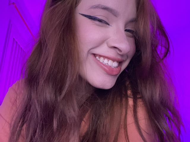 Madeleine, met lang bruin haar, zwarte eyeliner met een vleugvorm, lachend met roze lippen, waarbij witte tanden zichtbaar zijn, en een lichtroze huid, op een paars verlichte achtergrond..