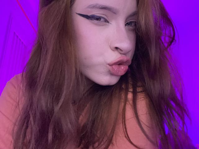 Madeleine: Eine Frau mit langen, braunen Haaren, schwarzem Eyeliner und vollen, rosafarbenen Lippen, die ein rosafarbenes Hemd trägt, hat einen Nasenring und ein violettes Hintergrundbild..