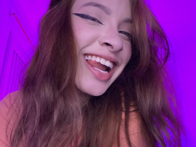 Madeleine, con lunghi capelli castani, eyeliner nero ad ali di gabbiano e un sorriso aperto e giocoso che mostra i denti bianchi, su uno sfondo viola. Il suo naso è leggermente all'insù..