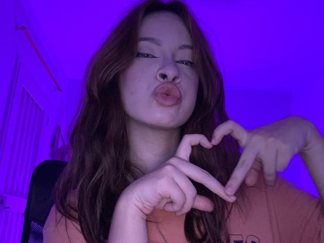 Madeleine, les lèvres pincées, les cheveux bruns ondulés, formant un cœur avec ses doigts, éclairage avec des nuances de violet, portant une chemise orange, petit anneau de nez légèrement visible..