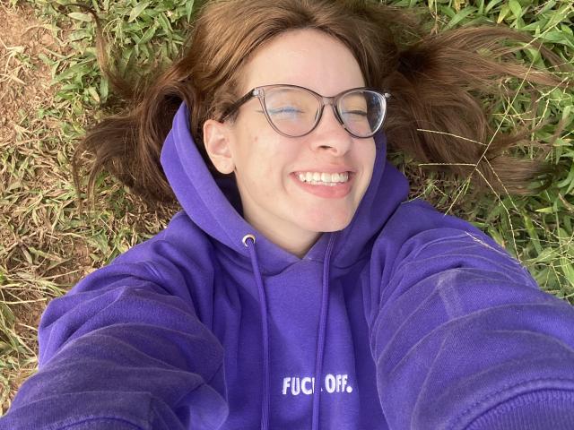 MadeleineBlack lying on grass, smiling, wearing a purple hoodie with "FUCK OFF" in white, and glasses, her brown hair splayed out, her tongue slightly out.