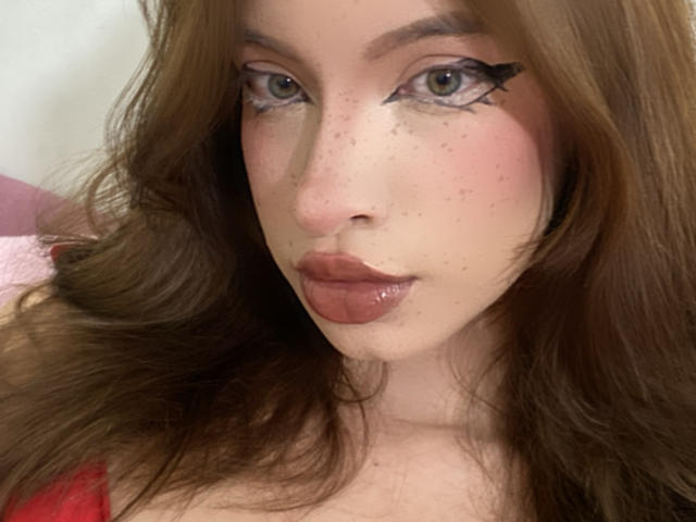 Madeleine: Zwarte lippen, bruin golvend haar, groene ogen, dikke zwarte eyeliner, sproeten op haar neus en glanzende roze lippen. Borsten gedeeltelijk zichtbaar in een rode top..
