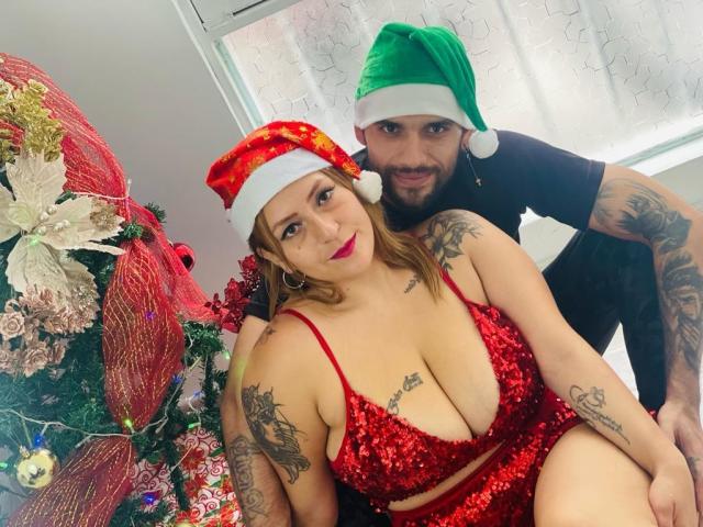 CloeAndDaniel met rode satijnen lingerie, grote borsten, tatoeages en een kerstmuts, zittend naast Daniel in een groene kerstmuts, beiden glimlachend, kerstboom op de achtergrond..