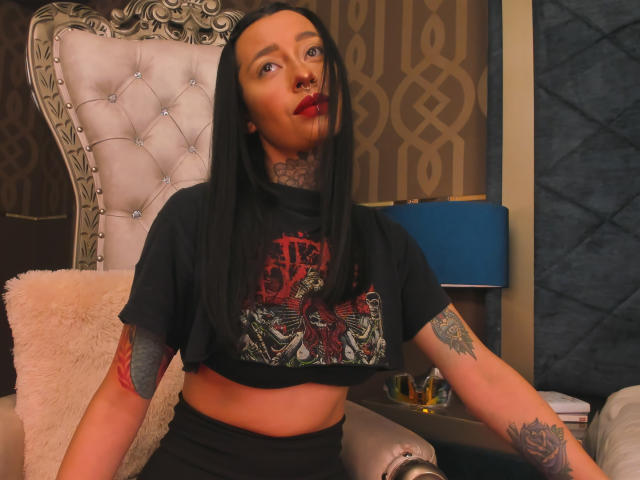 AlannaJade, con lunghi capelli neri, rossetto rosso, tatuaggi e un piercing al naso, indossa una maglietta nera con una grafica e pantaloni neri, seduta su una sfarzosa poltrona..