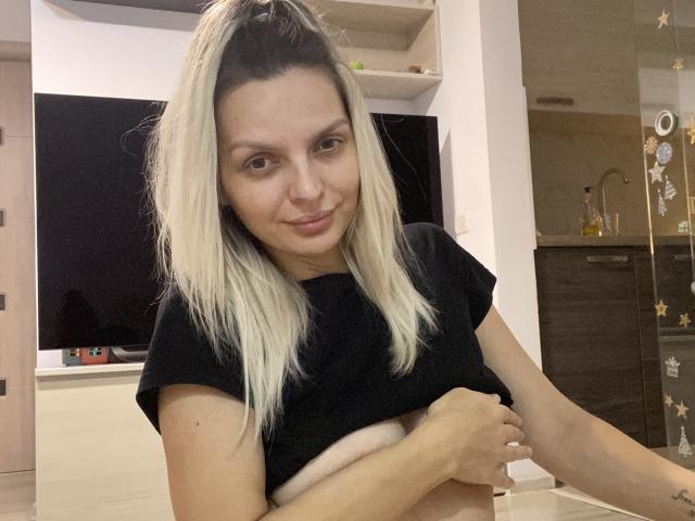 DouceAmelia with blonde hair, black t-shirt, and exposed tits, smiling slightly, holding her tits with one hand in a modern, dimly lit room.