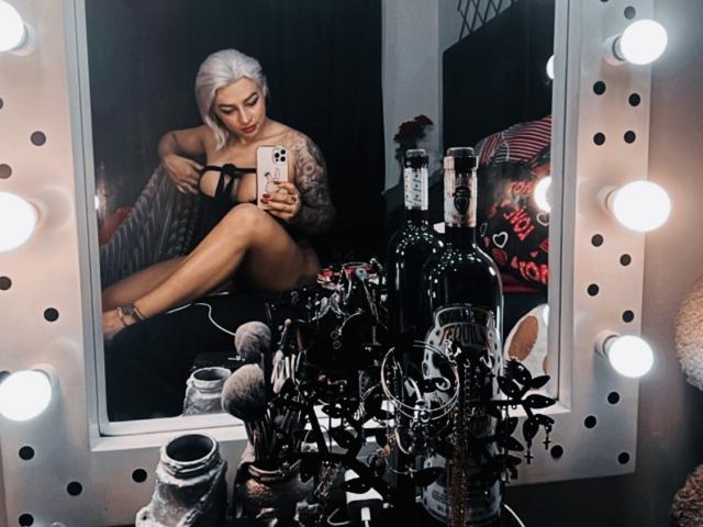 Avril Gray, com cabelo branco, tatuagens e vestido preto, sentada em frente a um espelho, segurando um telefone. O reflexo mostra ela tirando uma selfie. Há maquiagem e garrafas de bebida na superfície do espelho..