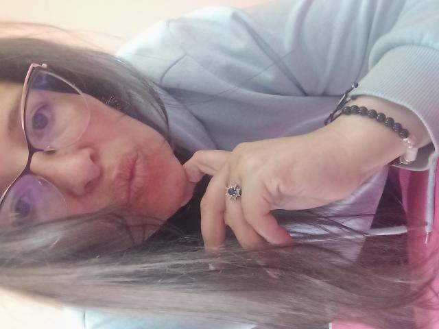 CandHoney with glasses, pouty lips, and long brown hair, wearing a light blue shirt, black bead bracelet, and a ring, looking into the camera.