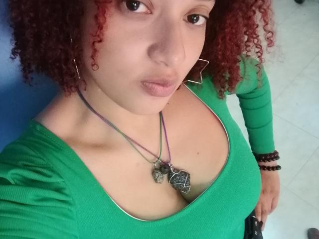 VioletaSwitch with curly red hair, green top, showing her tits, green eyes, pink lips, necklace with heart pendant, earrings, bracelet, standing on white tile floor.
