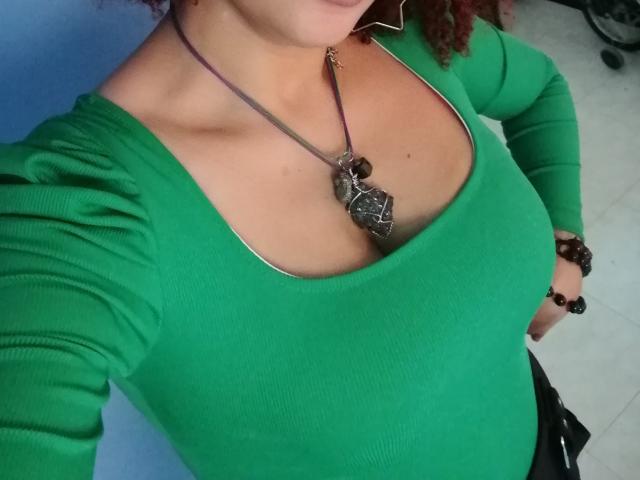 VioletaSwitch wearing a green ribbed shirt, necklace with a large stone pendant, and brown bracelet, showing her tits and part of her red curly hair.