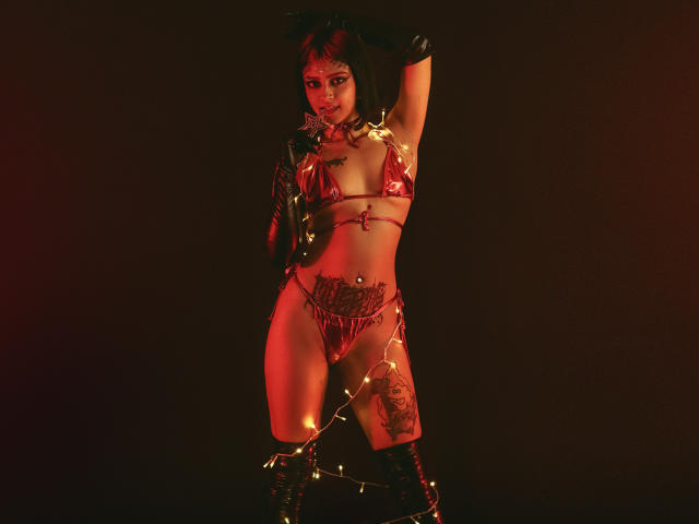 SerenaDeStark usando um biquíni vermelho brilhante, luvas pretas e botas, em pé com o braço levantado, com luzes de Natal brilhantes no corpo, tatuagens na barriga e na coxa, iluminação em vermelho escuro..
