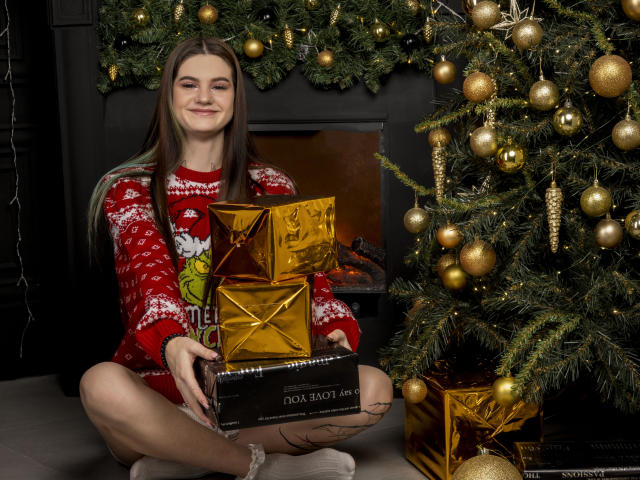 NicoleKissYou with long brown hair, wearing a red Christmas sweater, sitting cross-legged, holding gold and black award boxes, next to a decorated Christmas tree.