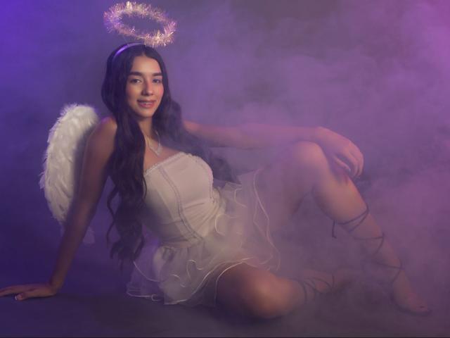 Caroliine Cossey, avec de longs cheveux noirs, un halo, une robe blanche et des ailes d'ange, assise par terre, souriante, les jambes croisées, éclairage violet et rose brumeux, avec des sangles sur ses jambes..