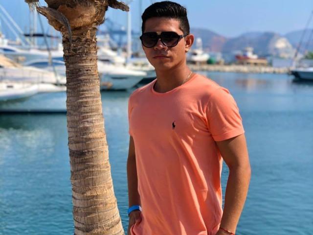 CalixHot está de pie junto a una palmera, vistiendo una camisa Lacoste de color rosa anaranjado, gafas de sol negras y una pulsera azul, frente a un puerto deportivo con varios barcos..