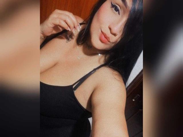 Briana69x, com cabelo escuro, vestindo uma regata preta, mostrando os seios, sorrindo, com uma tatuagem no ombro direito, segurando um colar, em um selfie..
