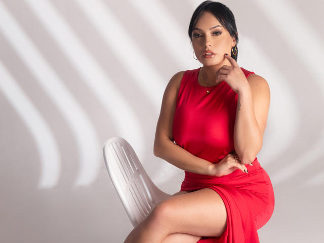 GalaMalkova, vestida con un ajustado vestido rojo, sentada en una silla blanca, tocándose la barbilla con una mano, mostrando su trasero bronceado y curvilíneo, y su cabello oscuro..