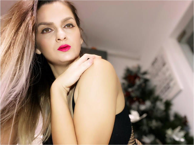 ReveMignoneX with long blonde hair, red lipstick, black tank top, and hand on shoulder, showing off her tits and cute face in a well-lit indoor setting.