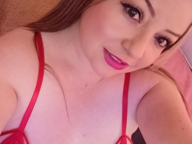 NatalyHami with pink lipstick, red bra straps, and long brown hair, smiling at the camera, showing her tits. Her eyes have black eyeliner and her skin is fair.