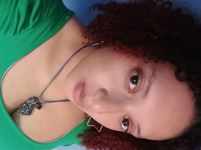 VioletaSwitch with curly red hair, green top, and necklace, looking up with slightly parted lips. Her tits are small and her ass is not visible.