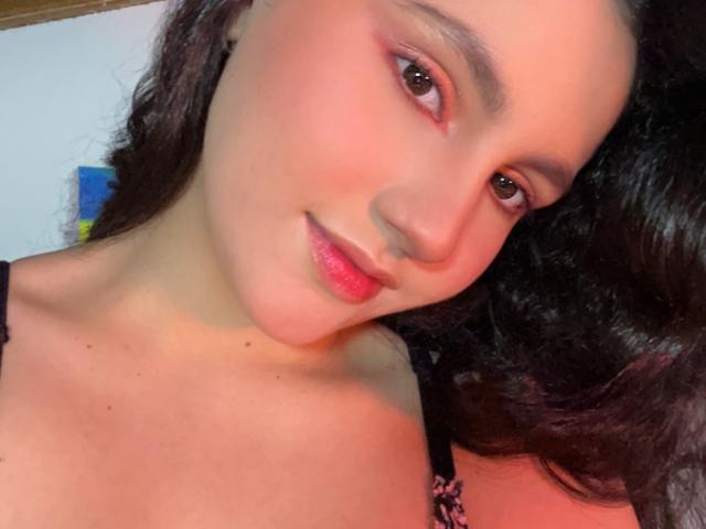 O rosto de EstefaniaRamosHot, com sombra rosa-avermelhada, lábios rosa brilhantes e cabelo escuro ondulado, olhando ligeiramente para o lado, vestindo uma blusa preta com alças. Iluminação suave e quente..