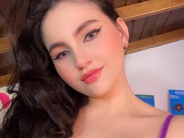 EstefaniaRamosHot heeft glanzende, roze lippen, zwarte eyeliner met een vleugje, en lang, zwart, golvend haar, met een schattige glimlach en een oorbel met een kleine ring, en draagt een paars top..