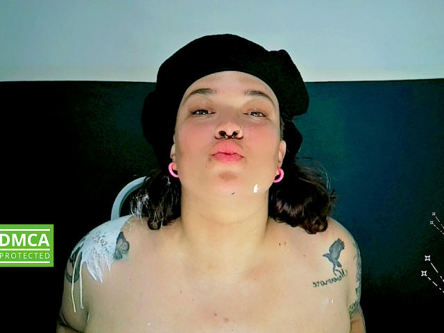 ElectricSapphire, con un berretto nero, auricolari rosa, tatuaggi, un anello al naso e labbra piene, mostra una goccia di liquido sulla guancia. È senza top, con tatuaggi visibili sulle spalle..