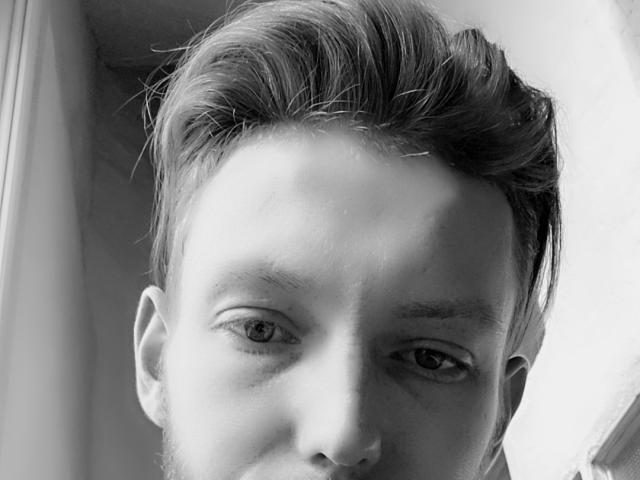 El rostro de Dylan Kyle en una fotografía en blanco y negro, que muestra sus ojos de color claro, su cabello ligeramente despeinado y una barba tenue, con una expresión tranquila y seria..